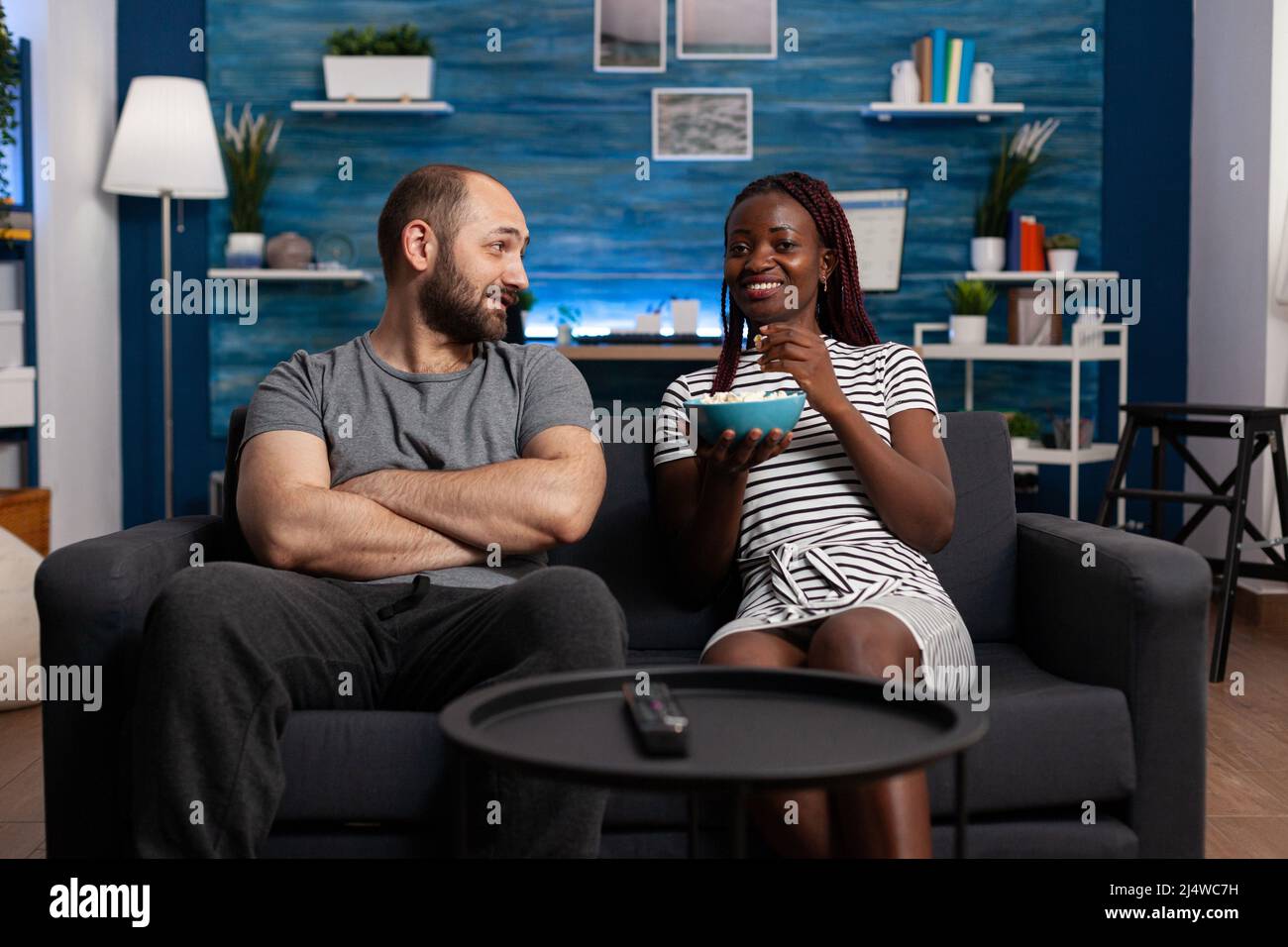 Happy interracial couple having a pleasent conversation while sitting on sofa at home. Relaxed man and woman bonding and having a good time in the living room while sharing snacks Stock Photo