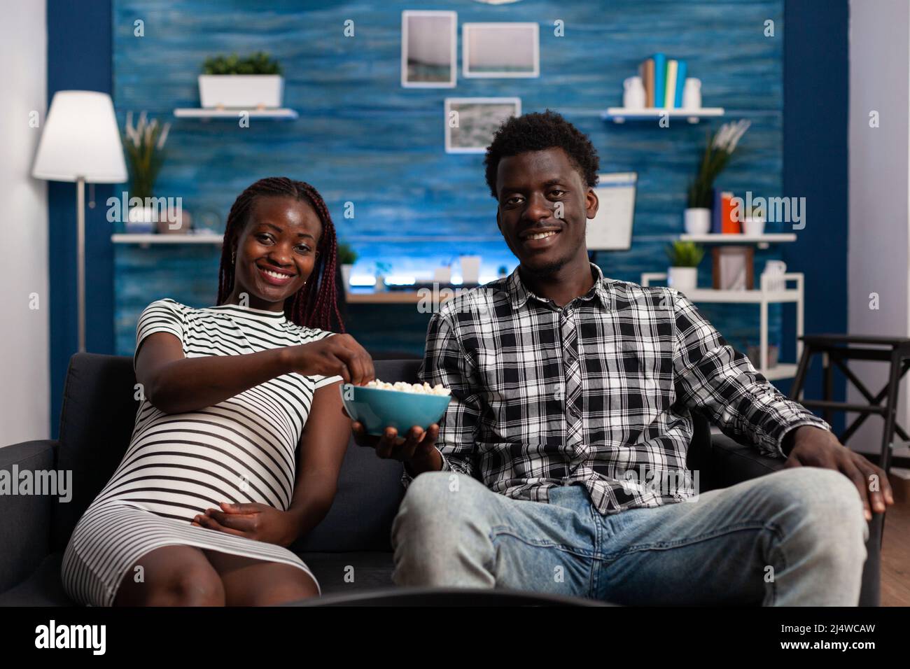 Smiling young african american couple sharing a bowl of popcorn while watching a television comedy program. Happy pregnant woman enjoying eating snacks sitting on couch while watching a movie in living room. Stock Photo