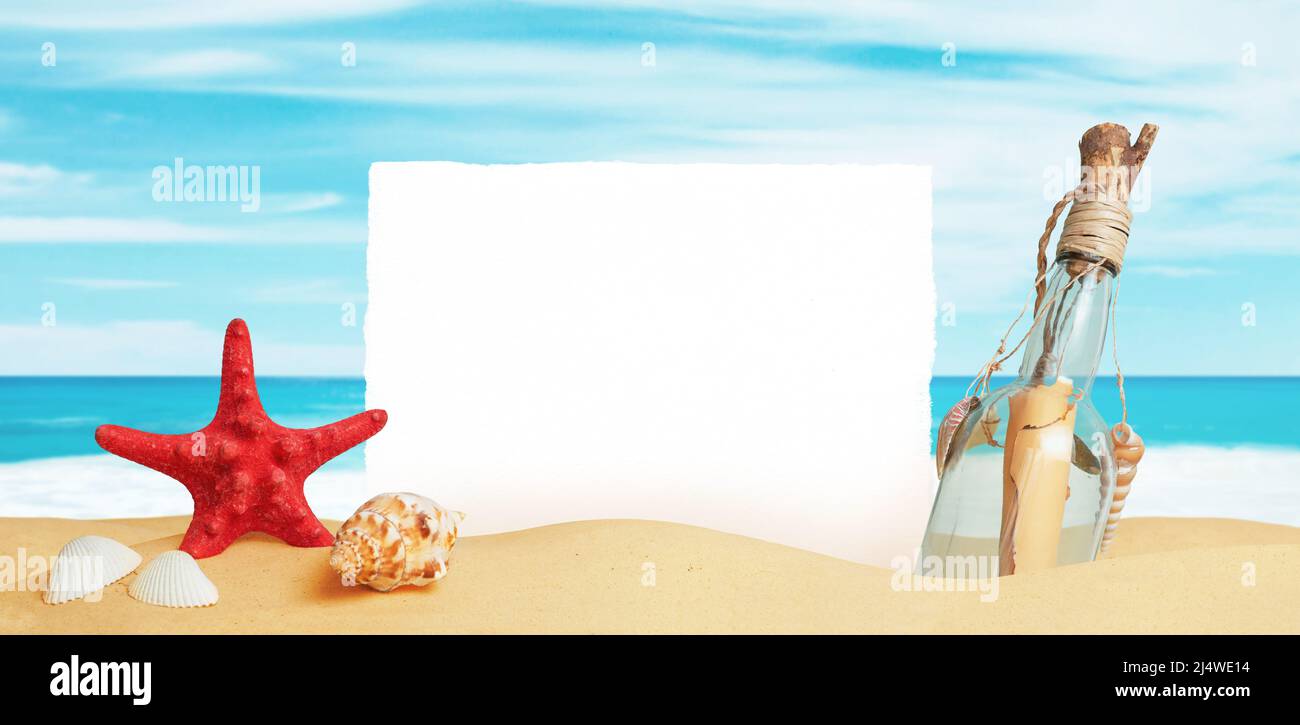 White paper stuck in the sand of the beach, surrounded by starfish, shells and a bottle with a message. Clean surface for promo text Stock Photo