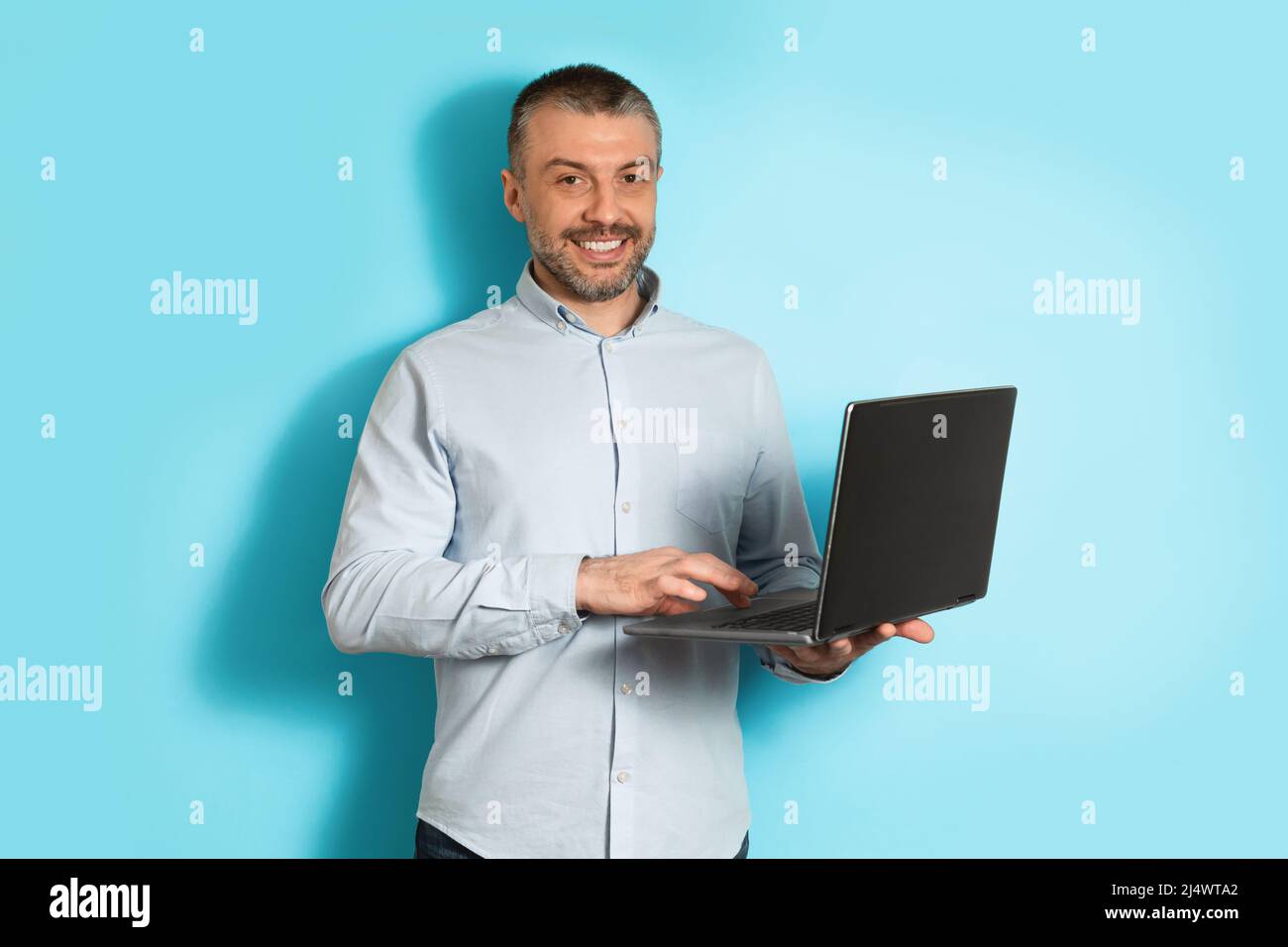 Middle Aged Man Using Laptop Computer Standing Over Blue Background Stock Photo