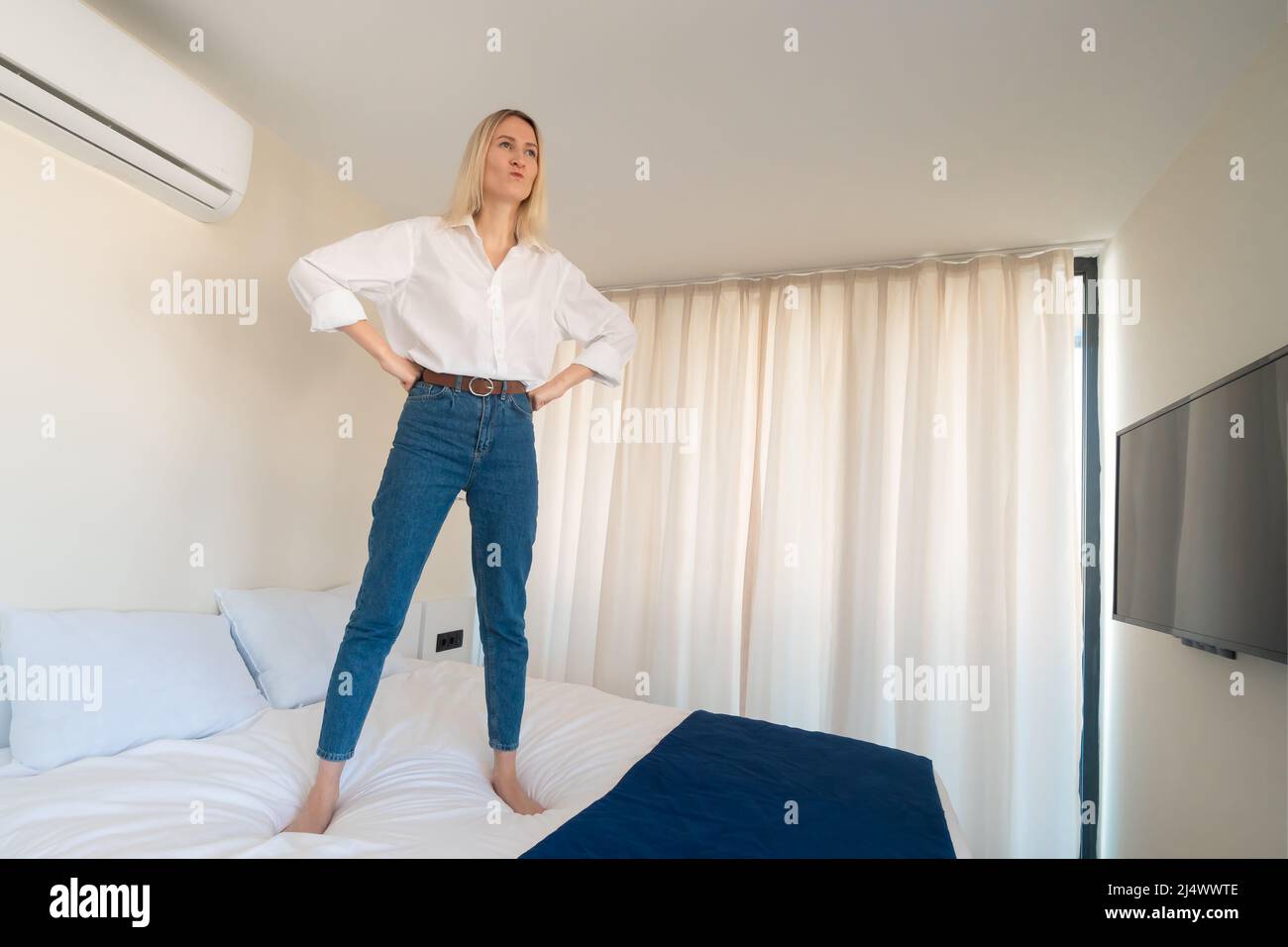 Moody, discontented young blonde woman with puffed cheeks in a white shirt, blue jeans is standing on the bed with her hands on her belt. Stock Photo