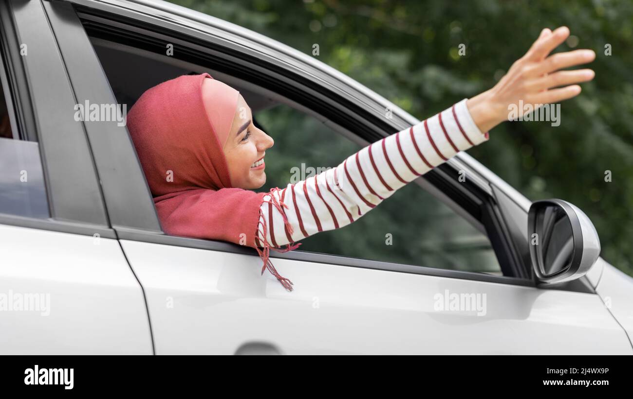 Satisfied millennial islamic woman in hijab catches air with her hand from open window in car, outdoor, panorama Stock Photo