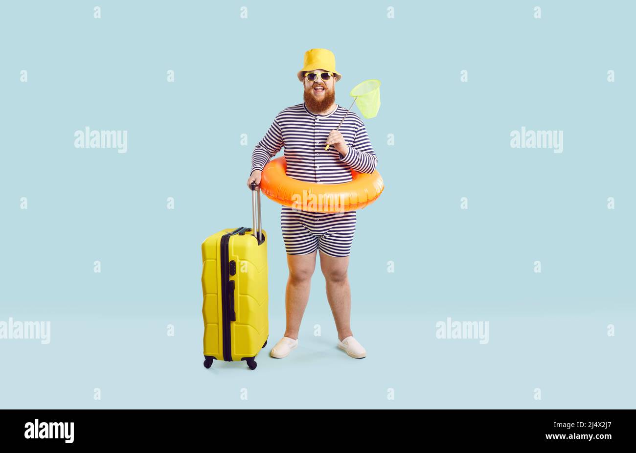 Funny fat man in swimsuit, hat, glasses and inflatable ring standing in studio with his suitcase Stock Photo
