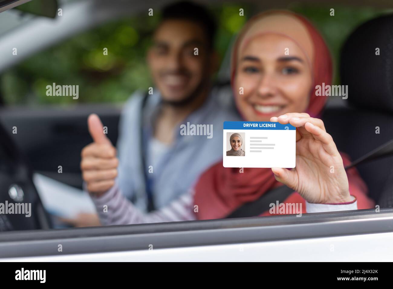 Glad middle eastern male and female in hijab show driving license and thumb up in open window in car after exam Stock Photo