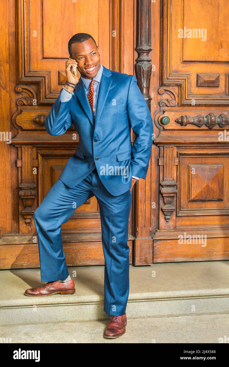 Dressing in blue suit, patterned shirt, necktie, short haircut, a young black businessman is standing by old style office door, feet stepping on stair Stock Photo