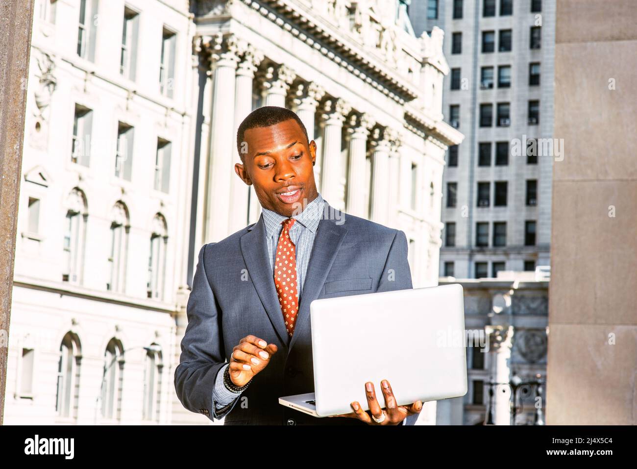Surprised businessman: a black man standing in the front of vintage style office building, looking down at laptop computer, raised eyebrows, opened ey Stock Photo