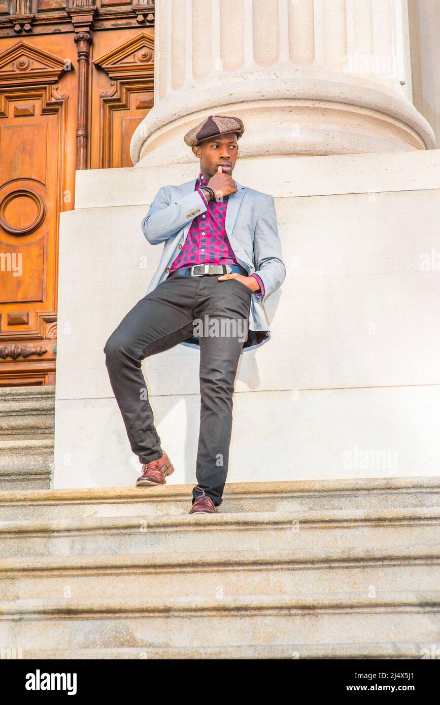Waiting for you. Wearing newsboy cap, dressing in blazer, pants, leather shoes, a young black guy standing on stairs outside vintage style office buil Stock Photo