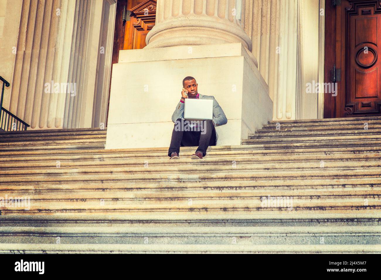 Way to Success. A black business man sitting on stairs of vintage style office building door way, working on laptop computer and talking on his mobile Stock Photo