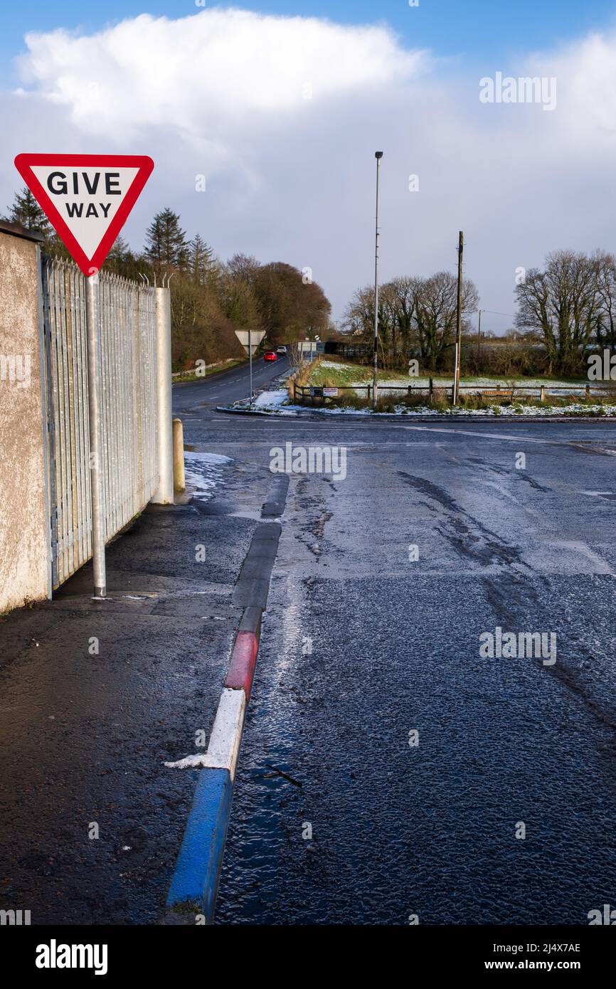 Red, white and blue painted kerb stones and a Give Way sign in the village of Ballybogey. In the distance a red car and a white car travelling away fr Stock Photo