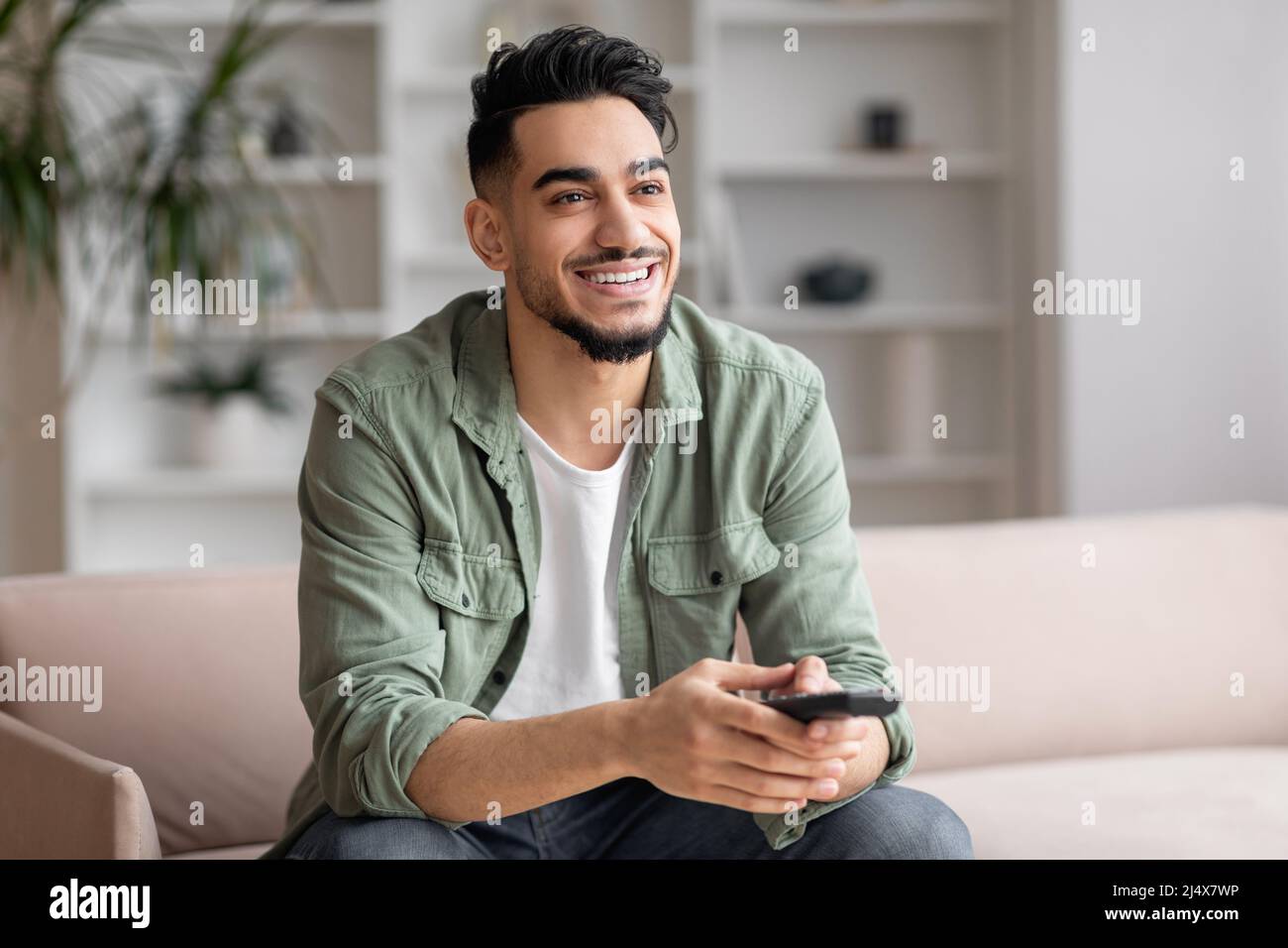 Cheerful handsome millennial arab man with beard in casual watching moving or match TV with remote control Stock Photo