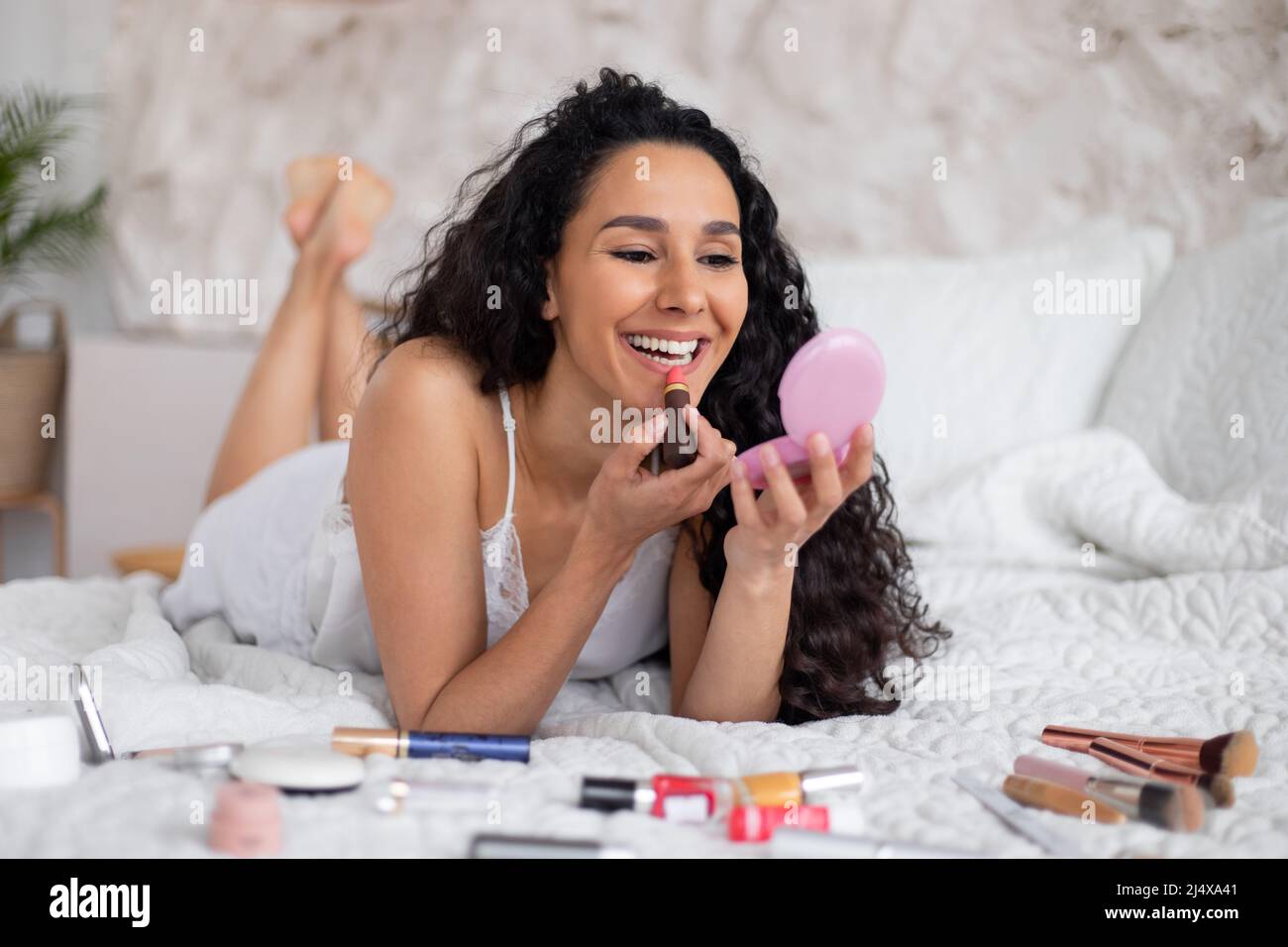 Cheerful young caucasian brunette woman with long curly hair in pajamas lies on bed and puts lipstick on lips Stock Photo
