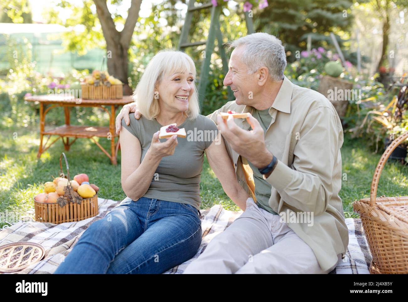 Happy aged couple having picnic in their garden, sitting on blanket and eating toasts with jam, enjoying spring day Stock Photo