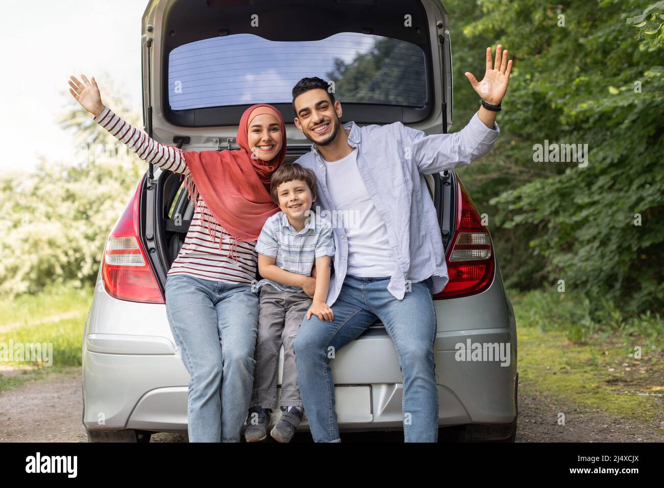 Satisfied millennial arab husband, wife in hijab and small boy waving hands sitting in car trunk Stock Photo