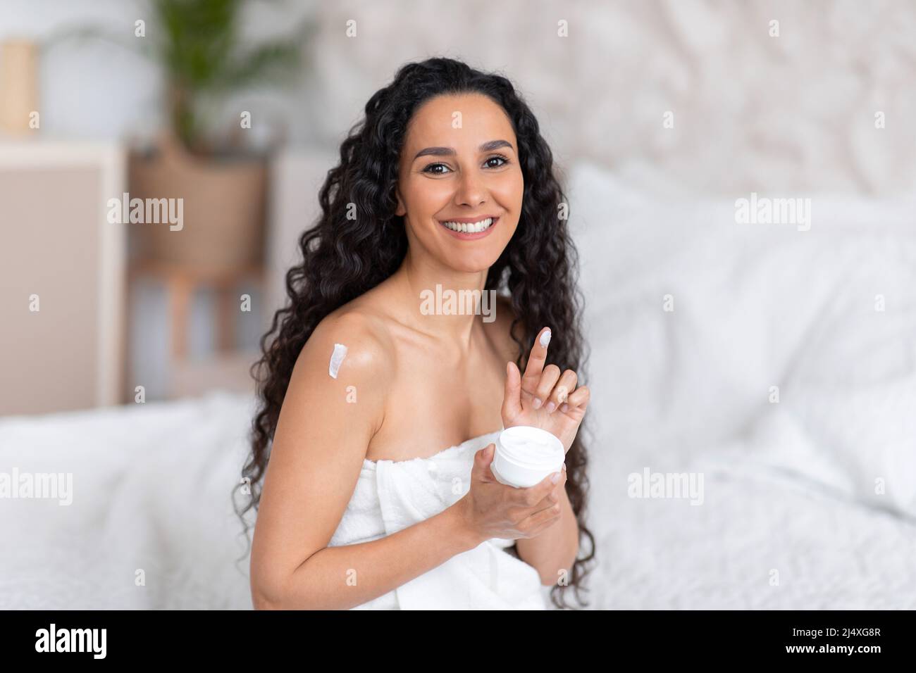 Cheerful millennial caucasian brunette female with long curly hair in towel applies cream on shoulder, hold jar Stock Photo