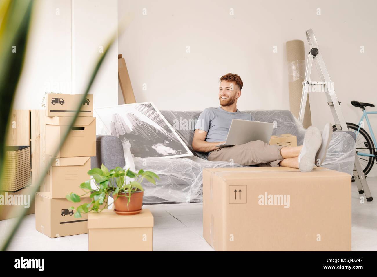 A relaxed young man sitting on the sofa using his laptop while smiling Stock Photo