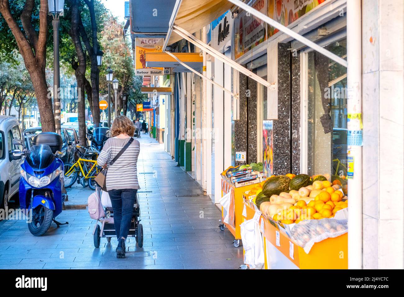 A woman pushes a stroller by a fresh fruits and vegetables store in the old town district. Valencia city is the capital of the Spanish province of the Stock Photo
