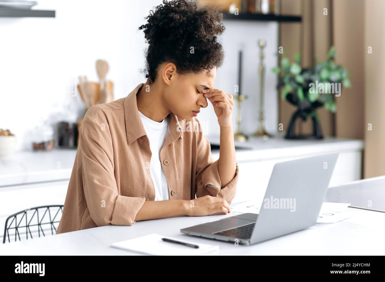 Overworked sad african american young woman, freelancer, student, trader, tired working using laptop, sitting at workplace at home, upset girl rubbing nose bridge, closed eyes, need break and rest Stock Photo