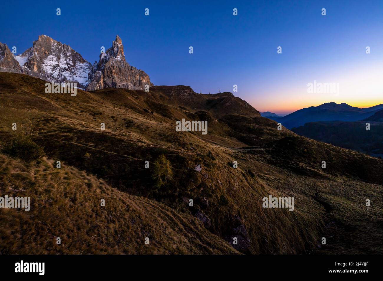 The Pala Group right after dusk, with a grass covered slope and a distant range on the horizon, as seen from Passo Rolle Stock Photo