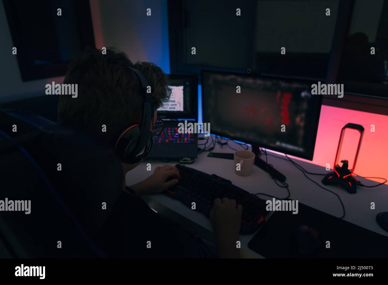 Young blond guy playing video games on a computer with headphones and using a backlit coloured keyboard. Stock Photo