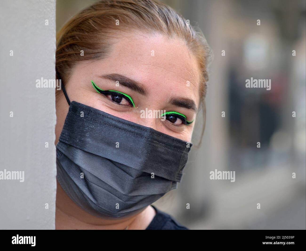 Young beautiful Mexican Latina woman with distinctive green eyeliner wears black surgical face mask and looks around a corner at viewer. Stock Photo