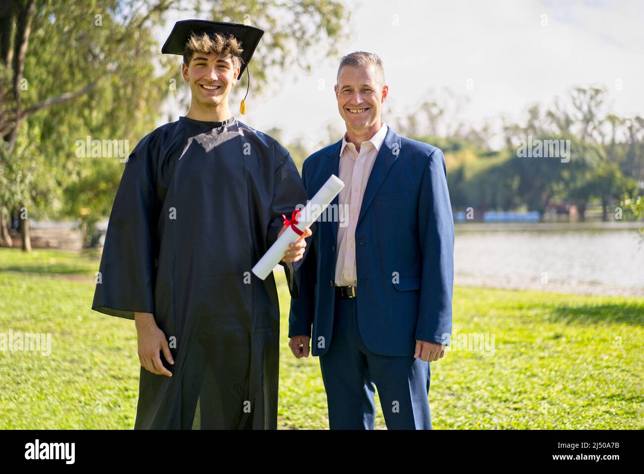 Young recently graduated boy, dressed in cap and gown, with his degree in hand, celebrating with his father on the university campus. Very happy expre Stock Photo