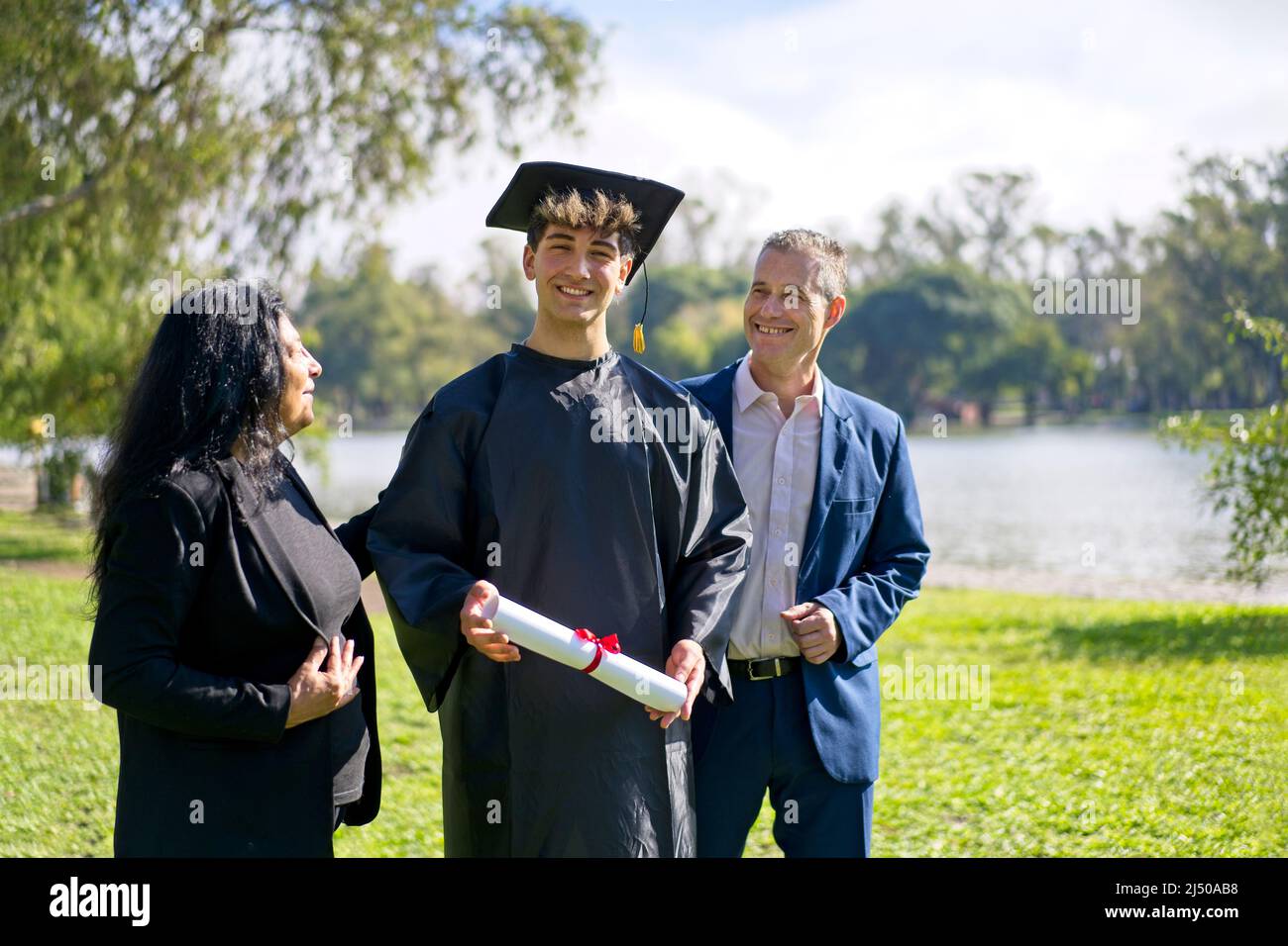 Young recently graduated boy, dressed in cap and gown, with his degree in hands, celebrating with his multi ethnic  family on the university campus. V Stock Photo