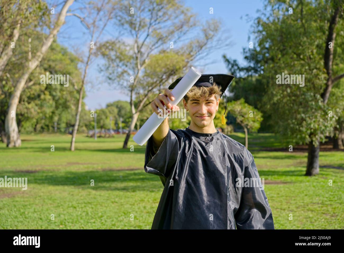Young recent graduate boy, dressed in cap and gown, showing off his degree, celebrating on campus at the university. Very happy expression, achievemen Stock Photo