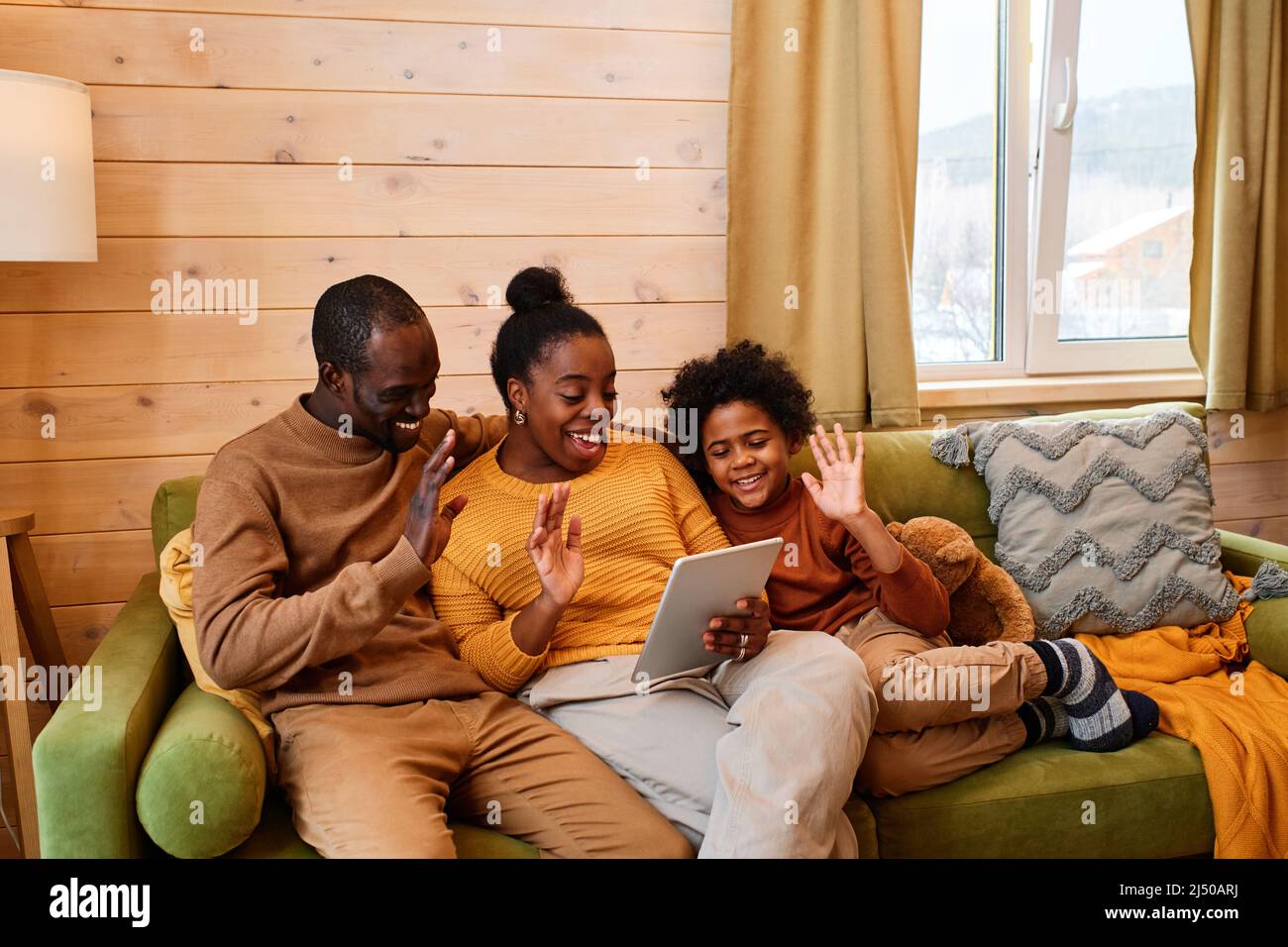 Happy family of three waving hands to someone on tablet screen during online communication while sitting on soft pistachio green couch Stock Photo