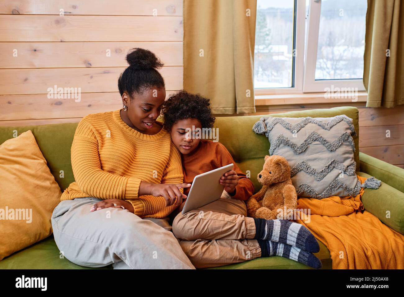 Young black woman in casualwear explaining something to her little son with teddybear while pointing at tablet screen at leisure Stock Photo