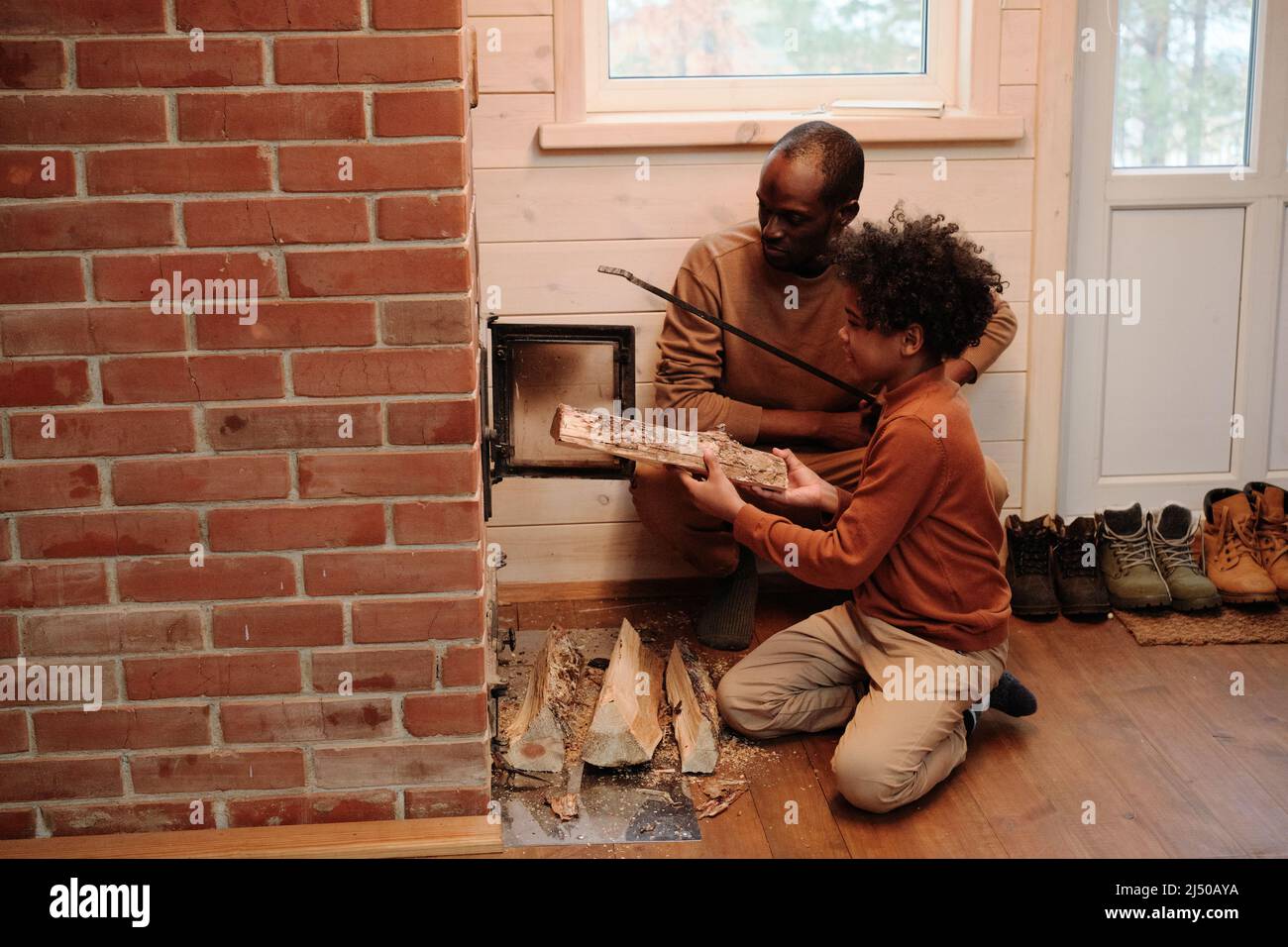 Cute little boy in casualwear helping his father fire up brick log burner to heat their country house while putting firewoods inside Stock Photo