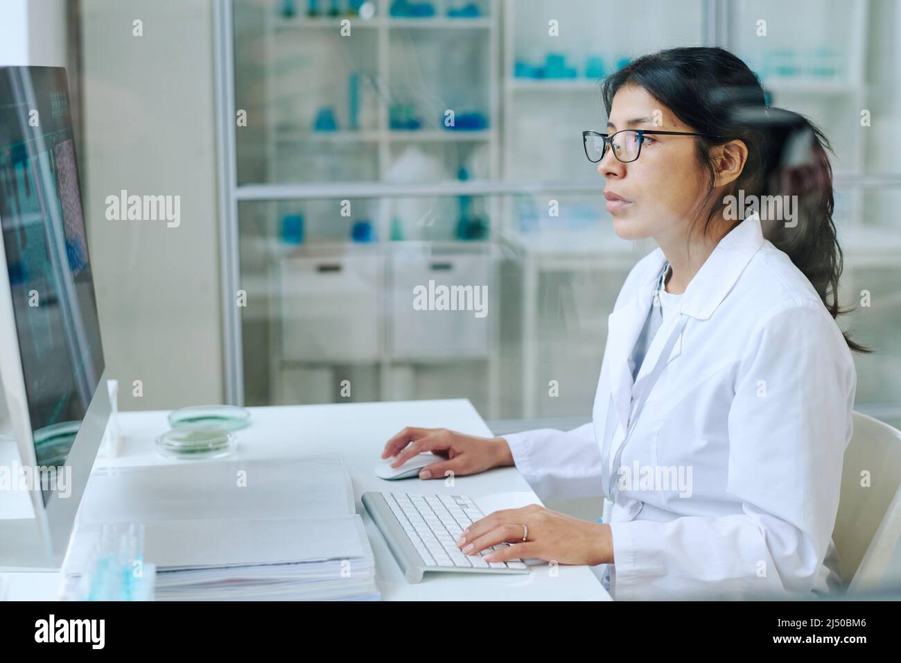 Young pretty female scientist in whitecoat and eyeglasses using computer for studying new virus while sitting by workplace in laboratory Stock Photo