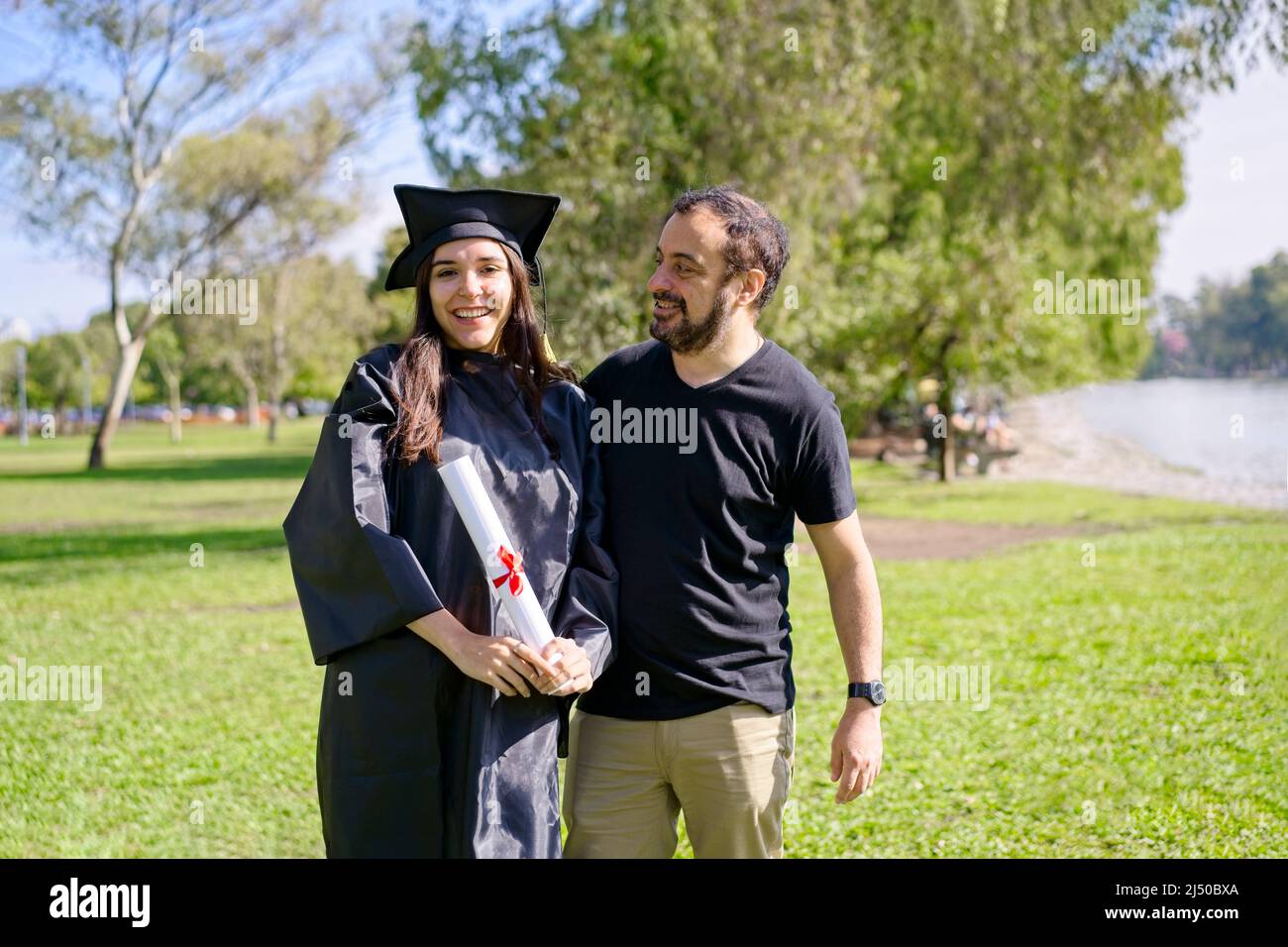 Young girl recently graduated, dressed in cap and gown, with her degree in her hands, celebrating with her family on the university campus. Very happy Stock Photo