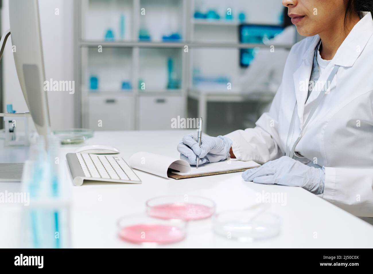 Gloved hands of young female scientist in whitecoat making notes in clinical document while sitting by workplace in laboratory Stock Photo
