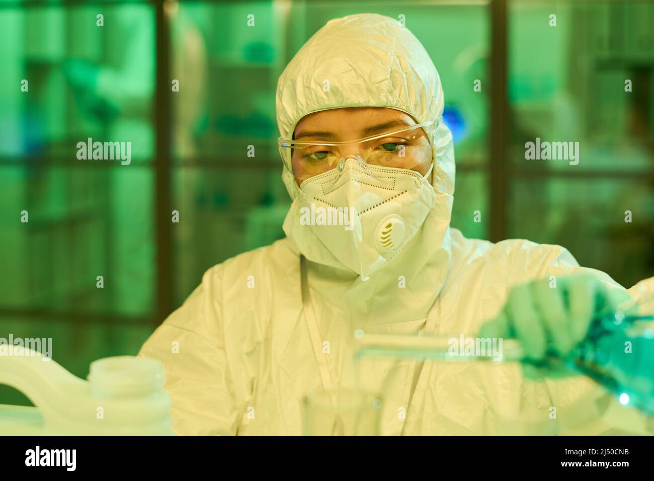 Young contemporary female scientist in protective coveralls, gloves and respirator pouring blue liquid from flask into other glassware Stock Photo