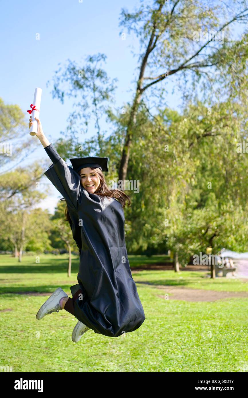 Young girl recently graduated, dressed in cap and gown, with her degree in her hand, jumping, on the university campus. Very happy expression, achieve Stock Photo