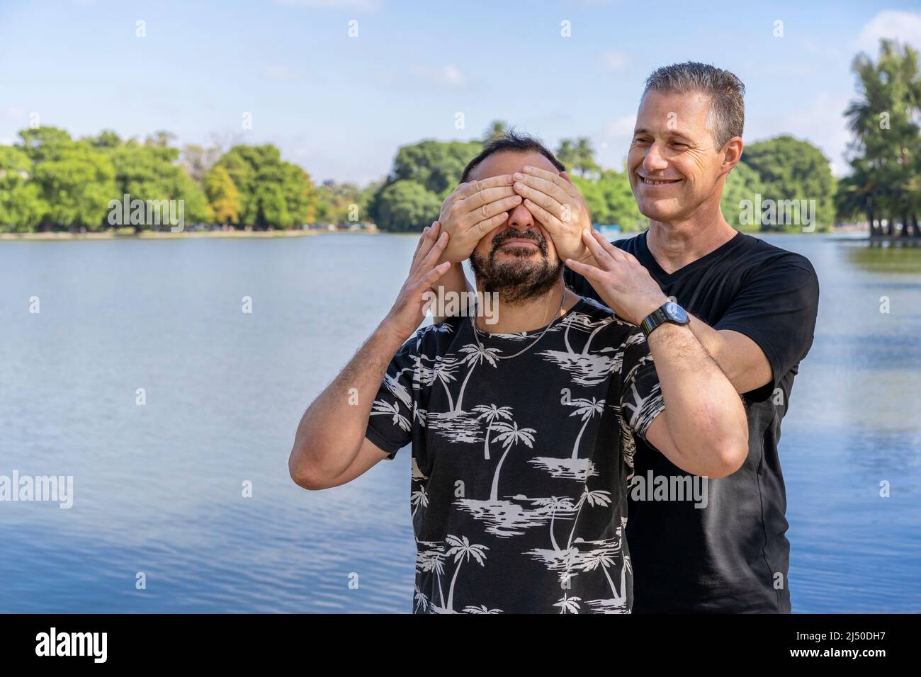 Couple of mature gay men, one surprising the other by covering his eyes in a lake. Concept of surprise, happiness, game Stock Photo