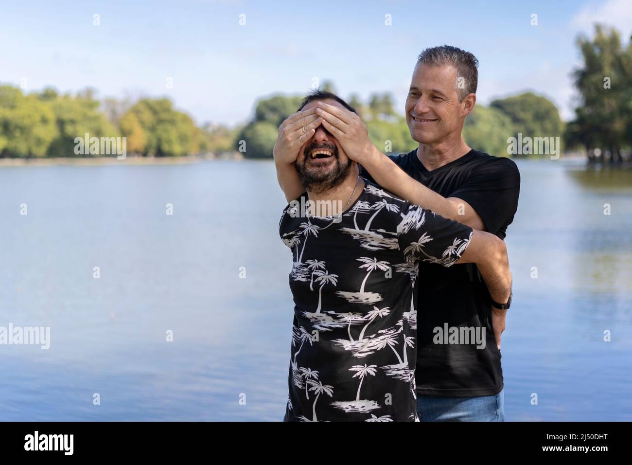 Couple of mature gay men, one surprising the other by covering his eyes in a lake. Concept of surprise, happiness, game Stock Photo