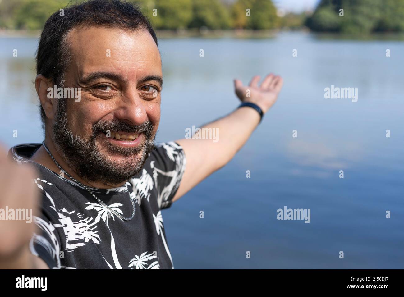 Active handsome mature Caucasian man taking selfie showing around at a lake. Outdoor adventure concept. Stock Photo