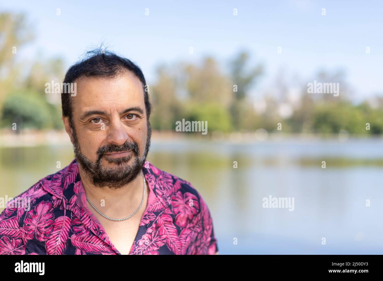 Bearded mature man wearing a fuchsia shirt, looking at the camera at a lake Stock Photo