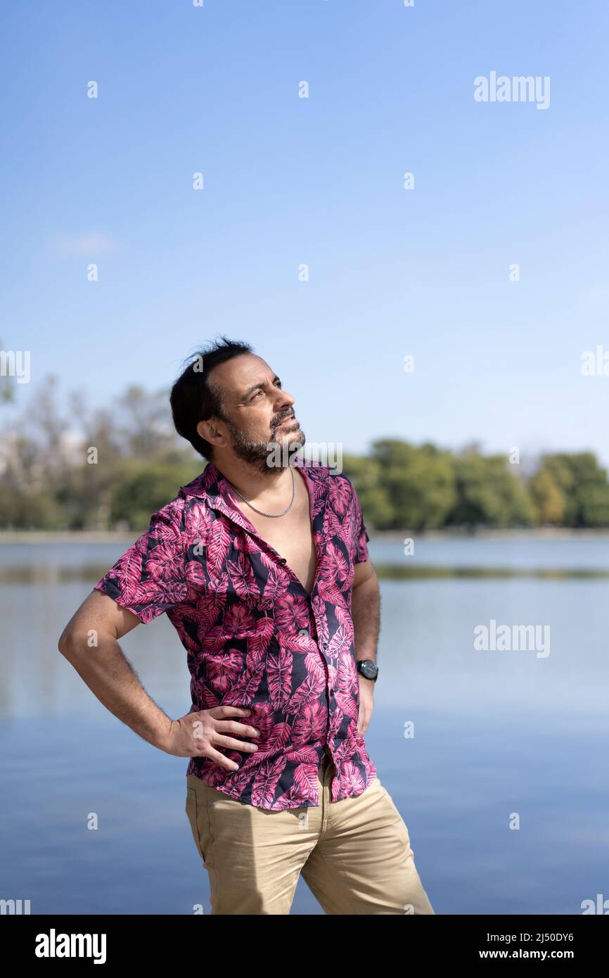 Bearded mature man wearing a fuchsia shirt, arms akimbo at a lake Stock Photo