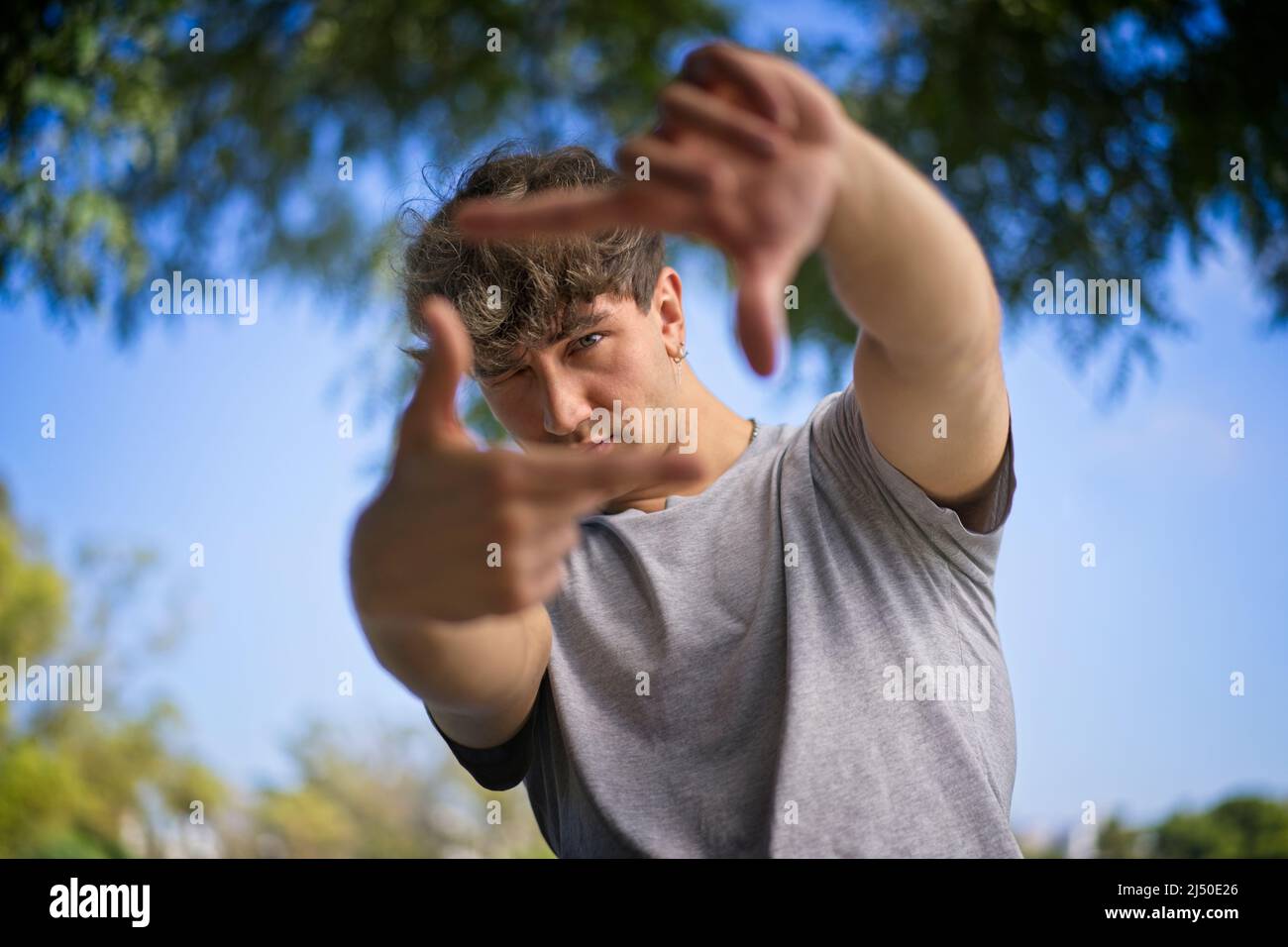 A handsome young man blinks his eyes and looks through a frame made up of hands. Attractive man make frame by fingers, isolated on blur background wit Stock Photo