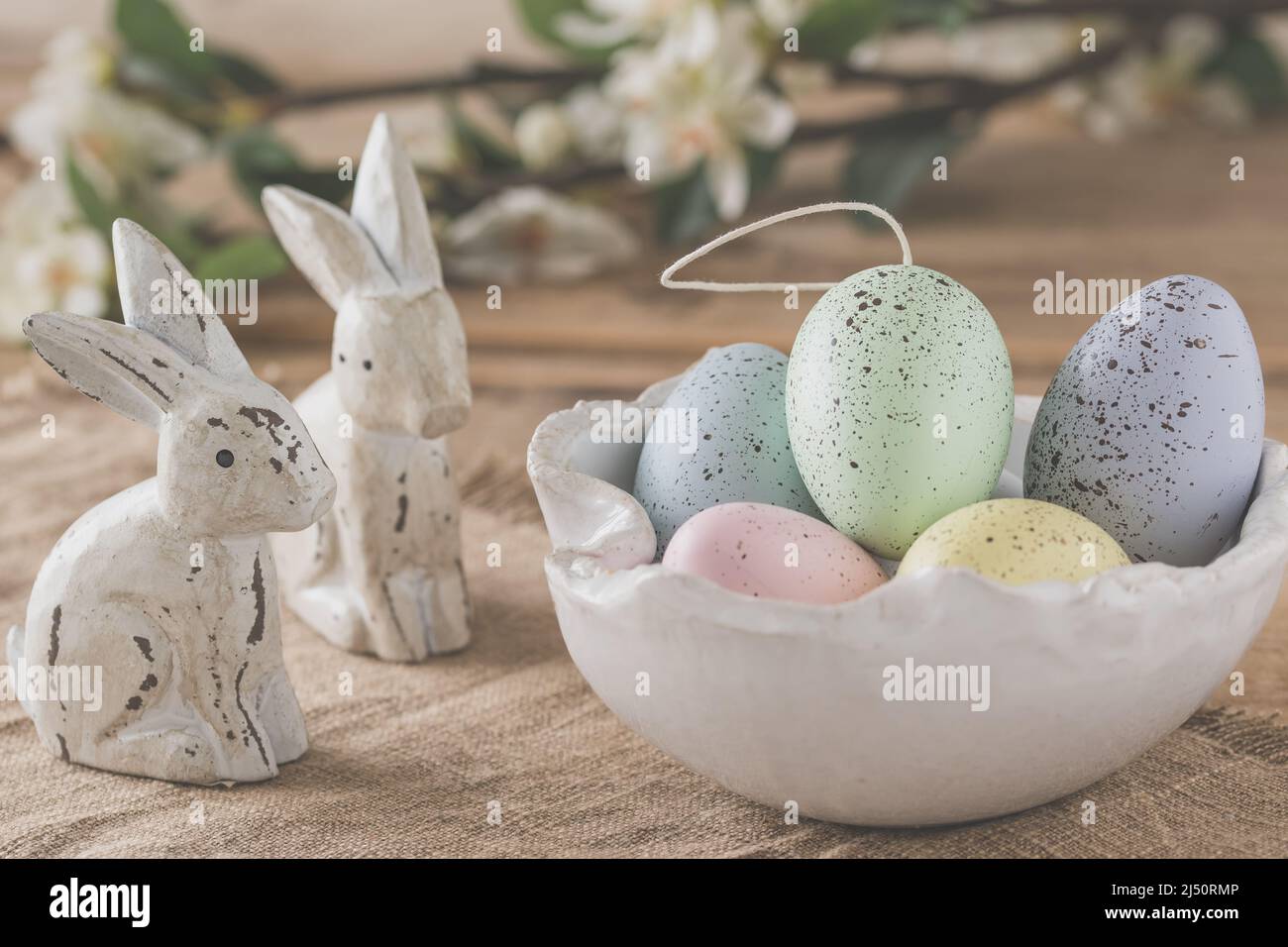 Decorative Easter eggs in pastel colors in a white handmade bowl and two wooden Easter bunnies on a wooden table, Easter decoration Stock Photo