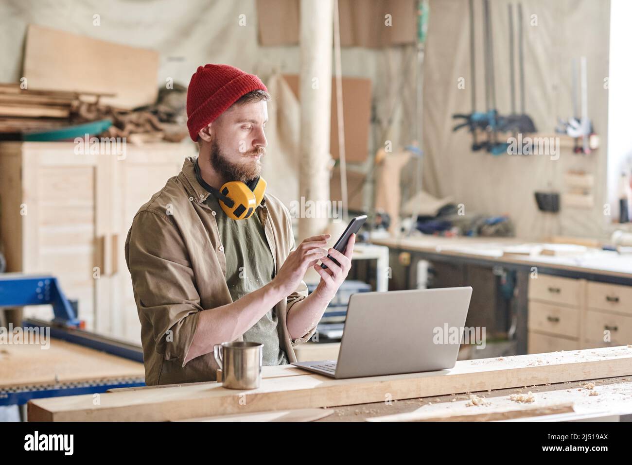 Modern woodworker standing at workbench with laptop and cup of tea on it texting something on smartphone Stock Photo