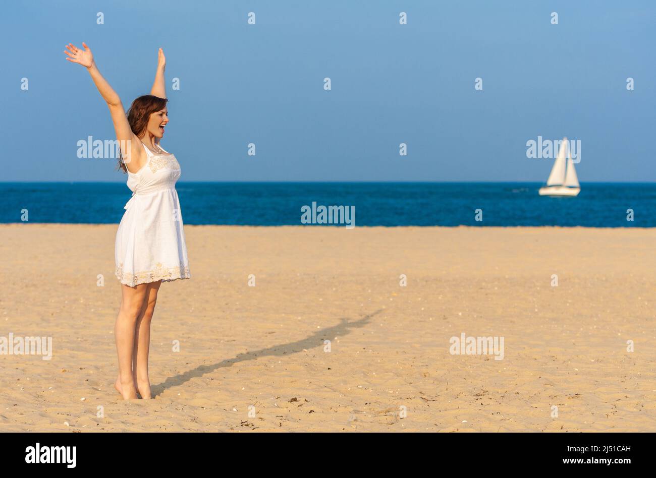 Young woman or girl standing on a beach celebrating arms raised on a beach with a white yacht or sail boat sailing at sea in background Stock Photo