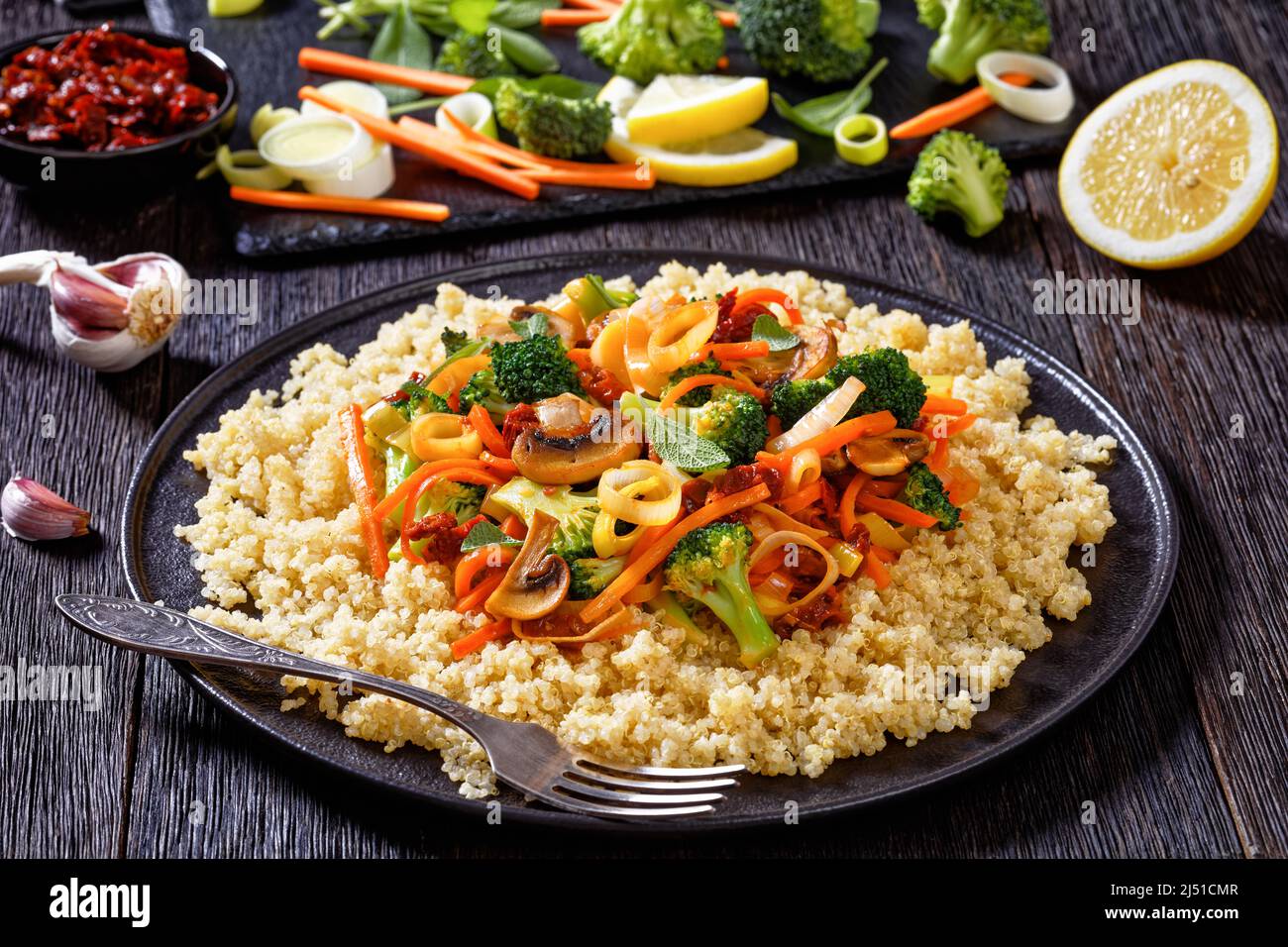 quinoa topped with stir fried broccoli, julienne carrots, sun dried tomatoes, leek and mushrooms on black plate on dark wooden table with ingredients Stock Photo
