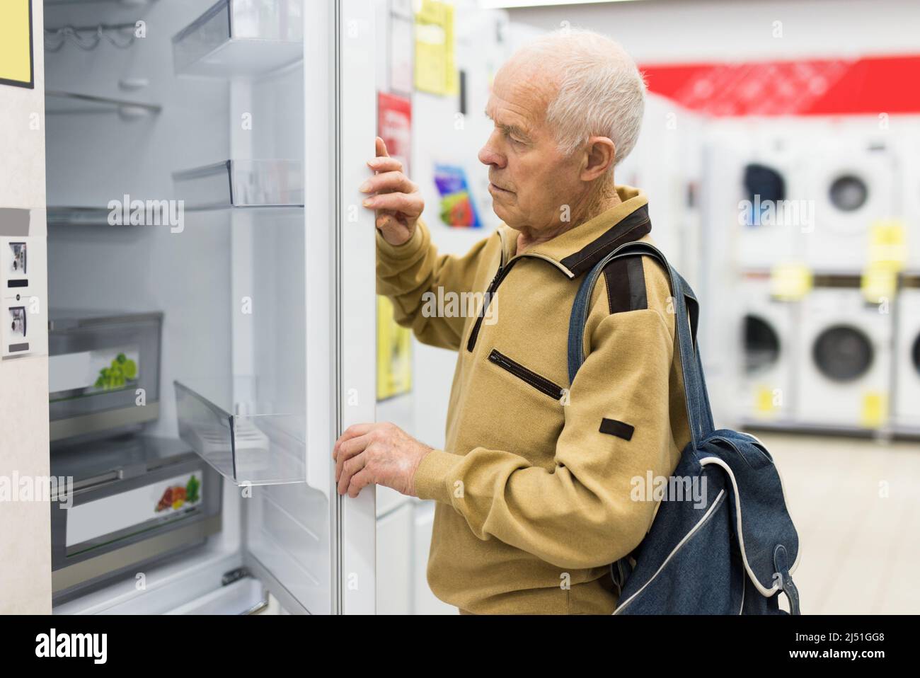 elderly grayhaired man pensioner looking refrigerator at counter in showroom of electrical appliance hypermarket department Stock Photo