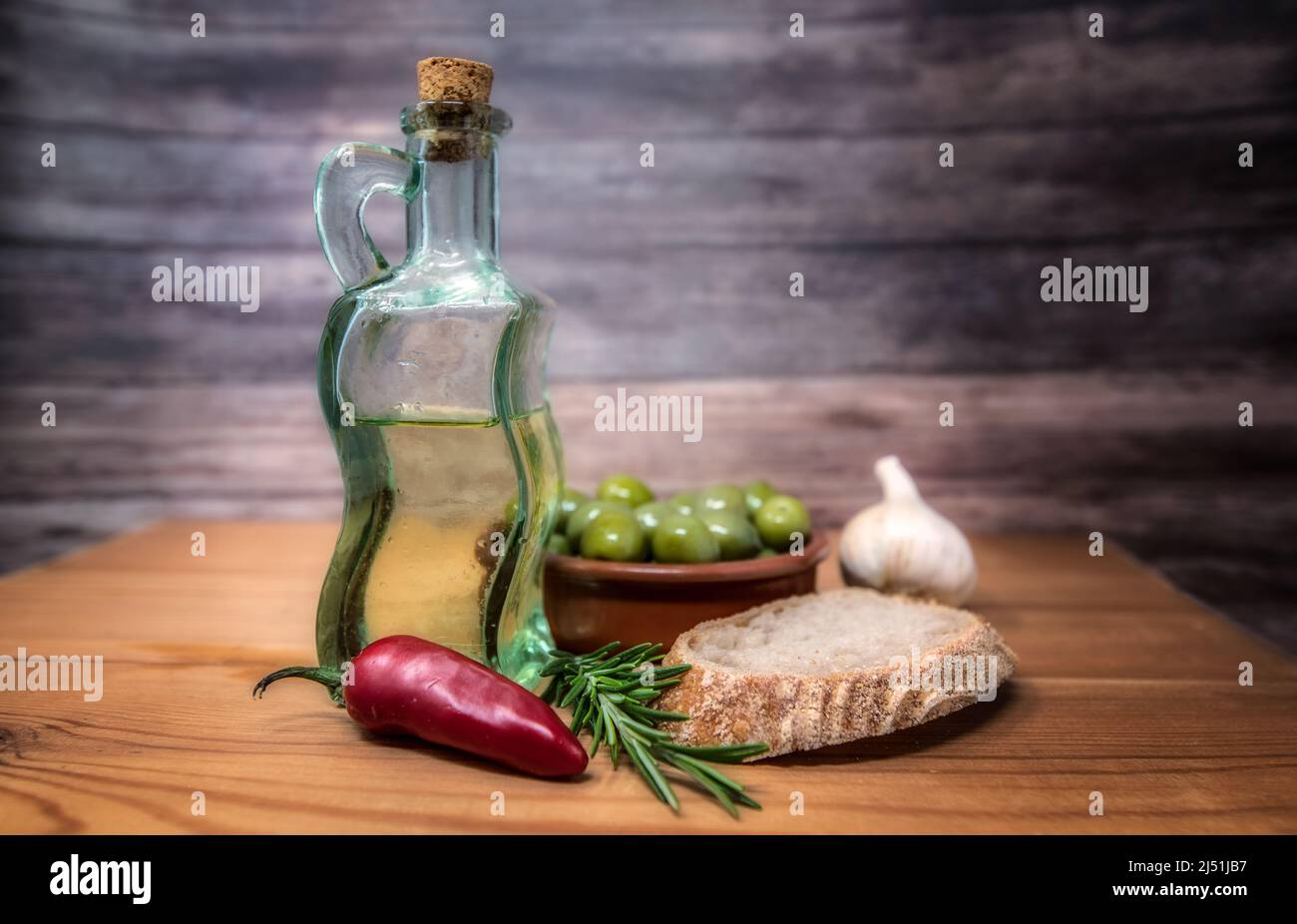 Table top still life of ovlive oil, green olives, red chilly, sprig of rosemary, garlic and bread in a rustic setting. Stock Photo