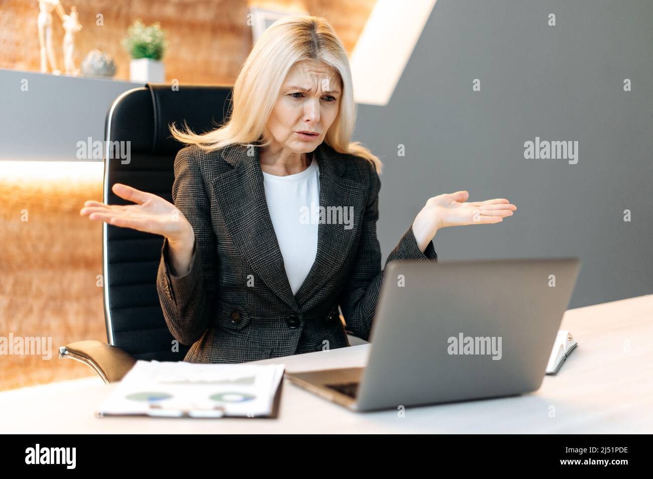 Discouraged upset middle aged blonde business woman, manager or broker, female executive, sit at the table in office, uses laptop, shocked by news or message, suffered financial loss,dissatisfied face Stock Photo