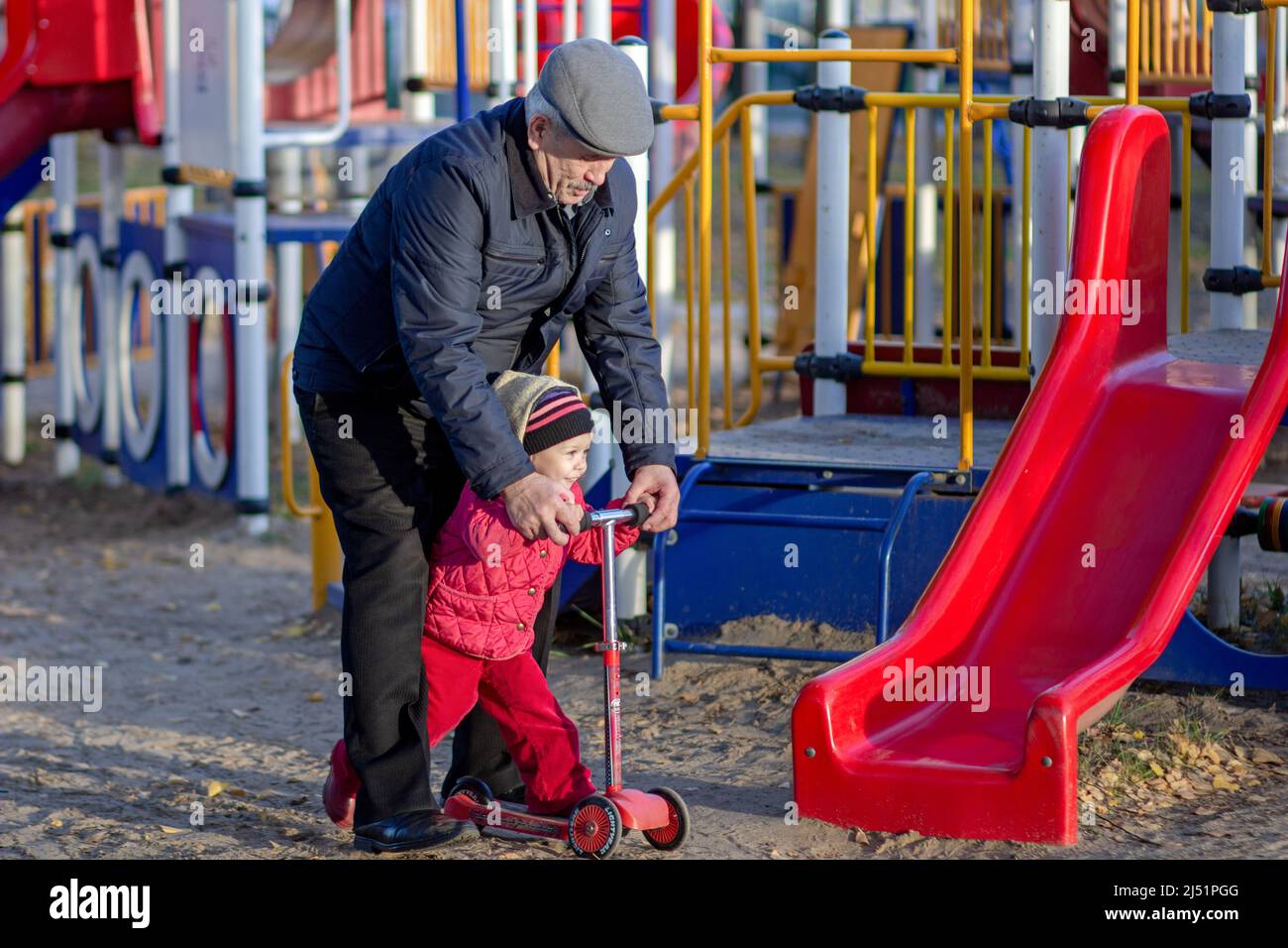 Caucasian grandfather helping granddaughter of 2-3 years to ride on scooter on playground outdoor Stock Photo