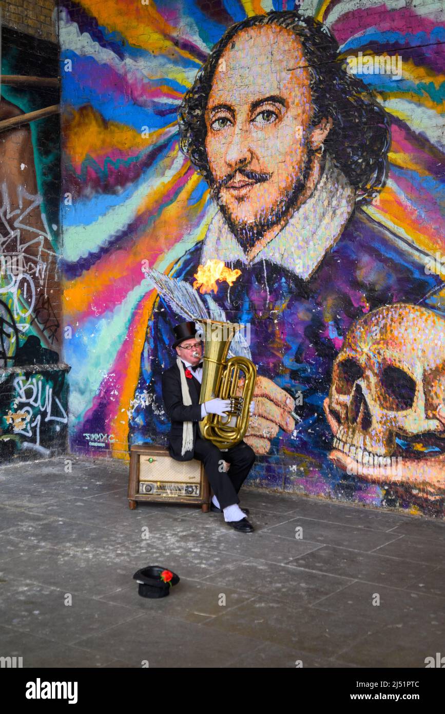 Busking tuba player breathing fire from his brass instrument under mural of William Shakespeare, Cannon Street Railway Bridge, South Bank, London, UK Stock Photo
